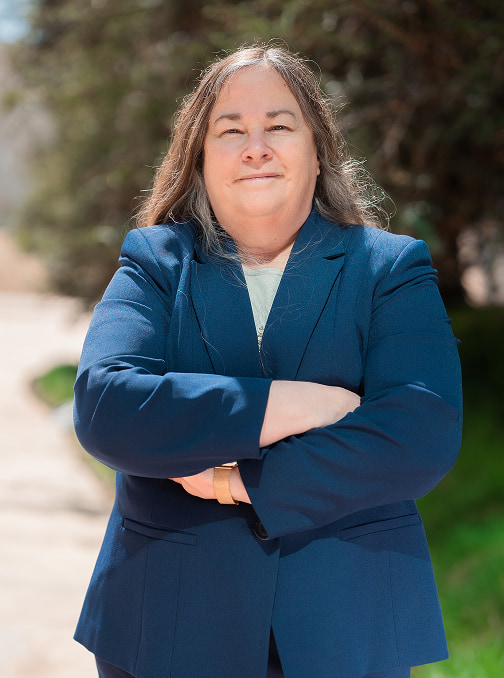 Profile photo of Caryn Sanders in a blue suit standing in front of nature background.