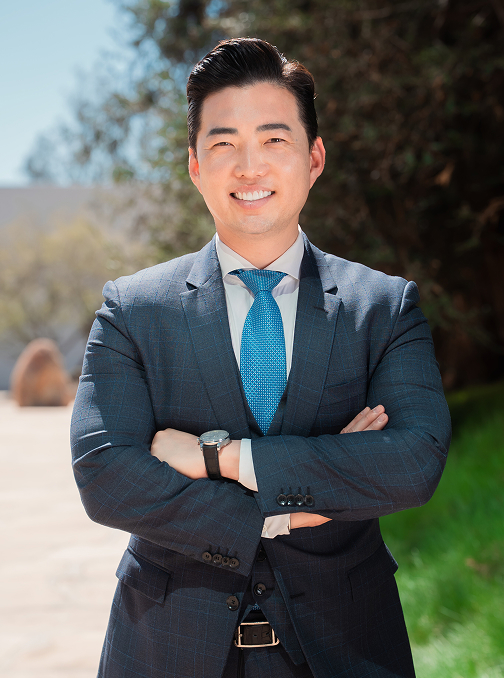 Individual in a blue suit and a blue tie with a green background and a tree behind them.