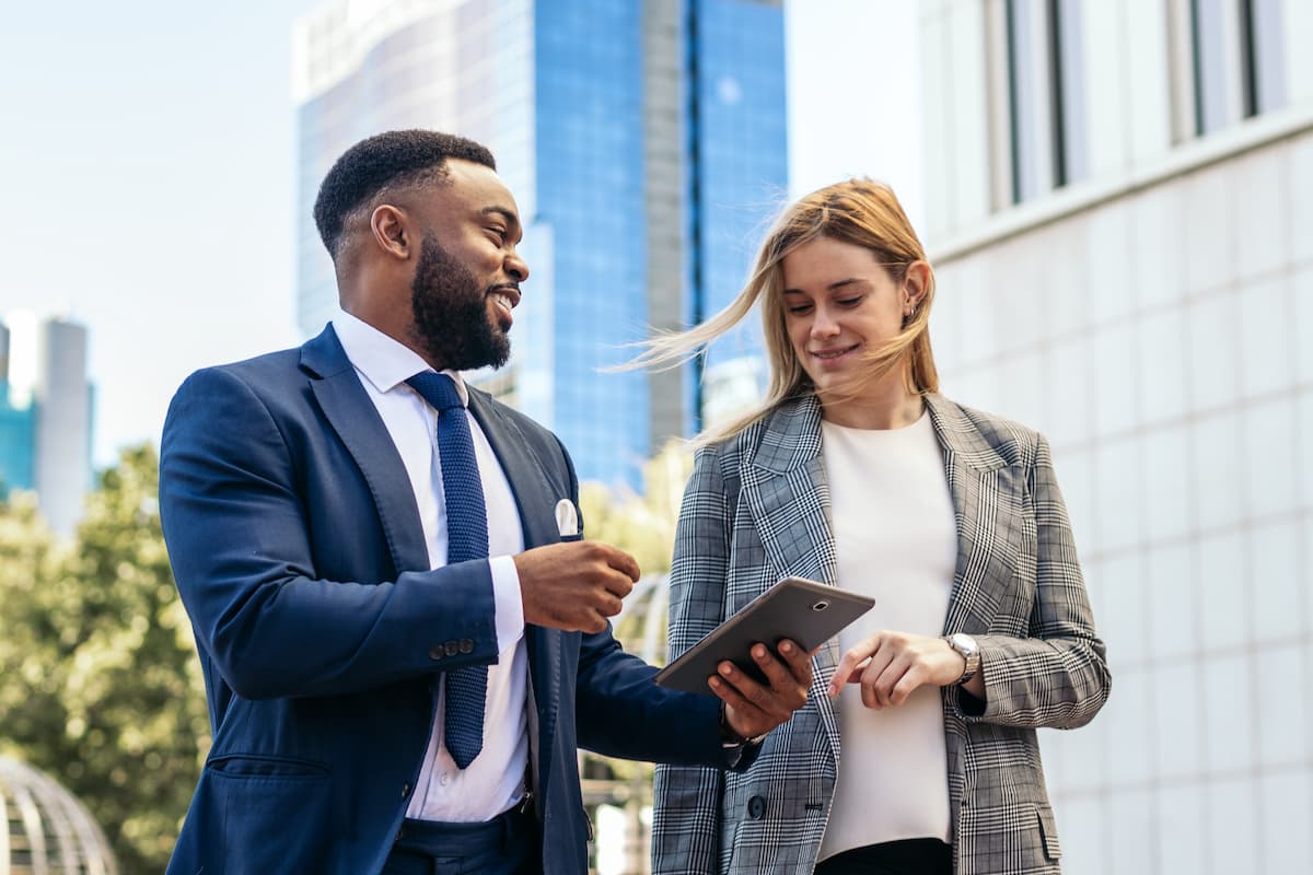Dos profesionales de los negocios, un hombre con traje azul marino y una mujer con americana a cuadros, caminan al aire libre en un entorno urbano, sonríen y comentan contenidos en una tableta digital.
