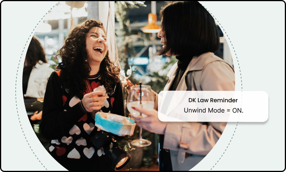 Two women holding drinks and laughing at a social gathering, with text overlay reading 'DK Law Reminder, Unwind Mode = ON.
