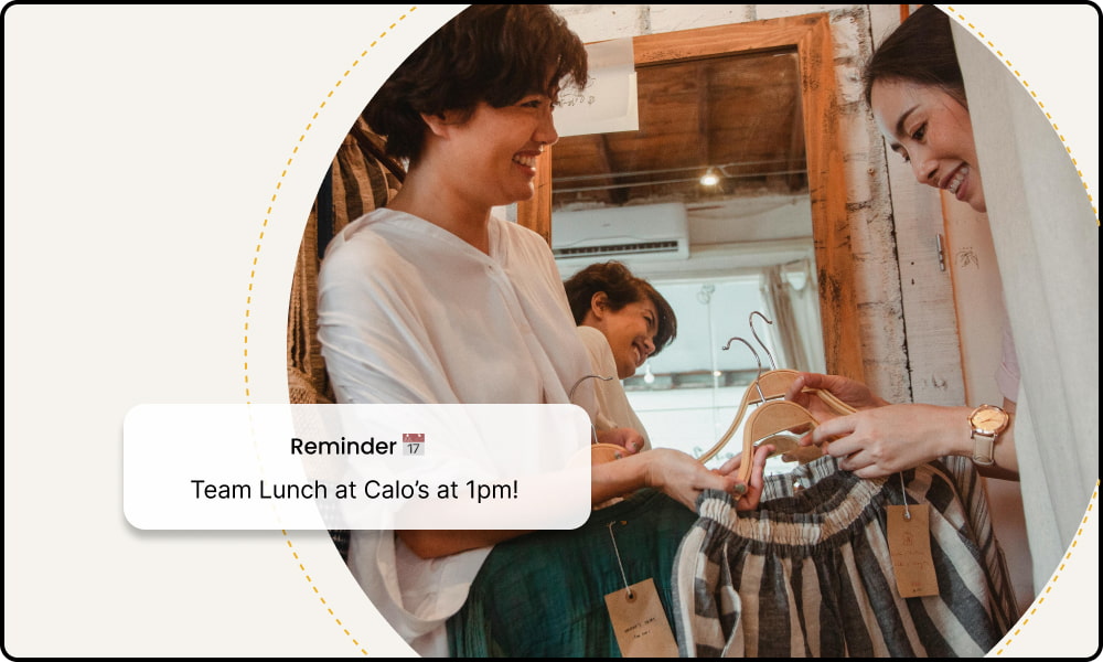 Two women smiling while holding clothes on hangers, with text overlay reading 'Reminder: Team Lunch at Calo’s at 1pm!