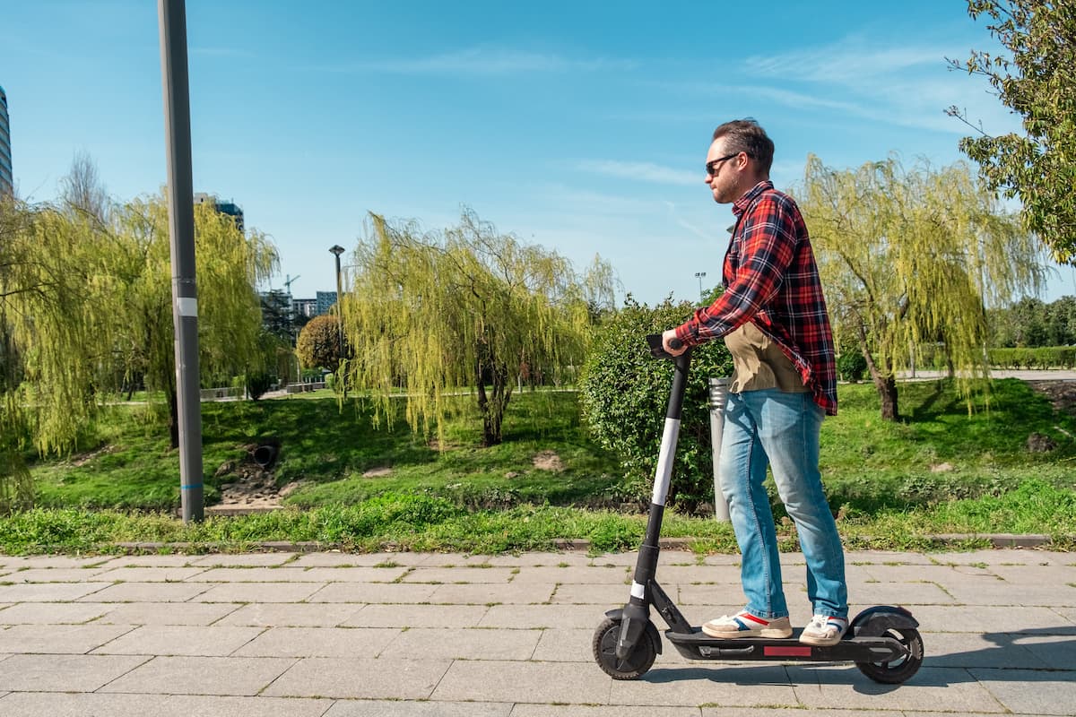 Side view of man riding e-scooter on pavement surface in city public park at sunny summer day.
