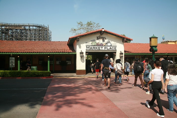 Entrance to Knott's Berry Farm Market Place in Buena Park, California. A group of people is walking toward the archway entrance under a sign with the Knott's logo and the words 'Market Place.' A wooden roller coaster is visible in the background on the left.