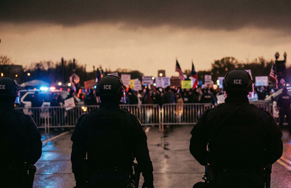 agents standing on the one side of a gate barrier. On the other side, people carry signs and American flags
