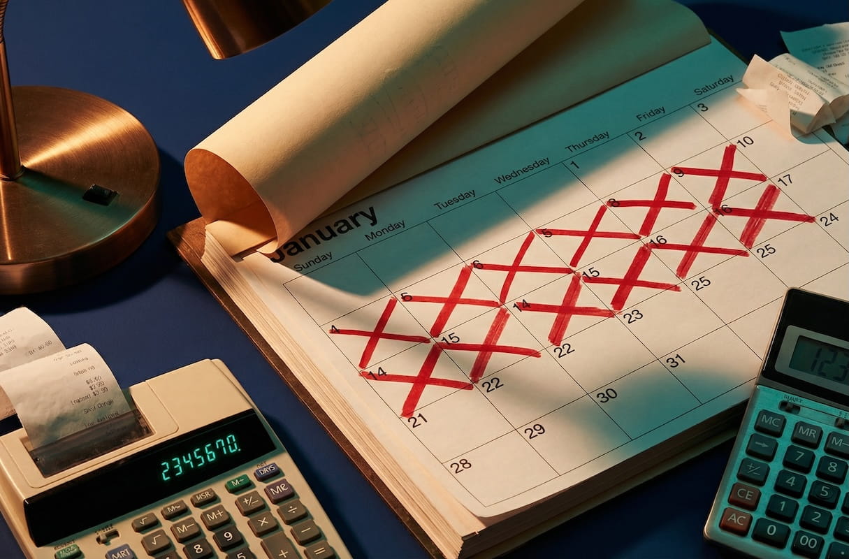 A January calendar with multiple days crossed out in red marker, surrounded by two calculators and receipts on a blue surface, symbolizing missed work days and lost wages following an accident.