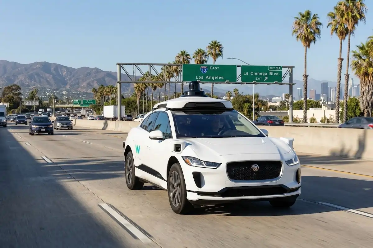 Waymo autonomous vehicle driving on Los Angeles freeway with palm trees and mountains in background. Self-driving car technology on Interstate 10 heading east toward downtown LA skyline.