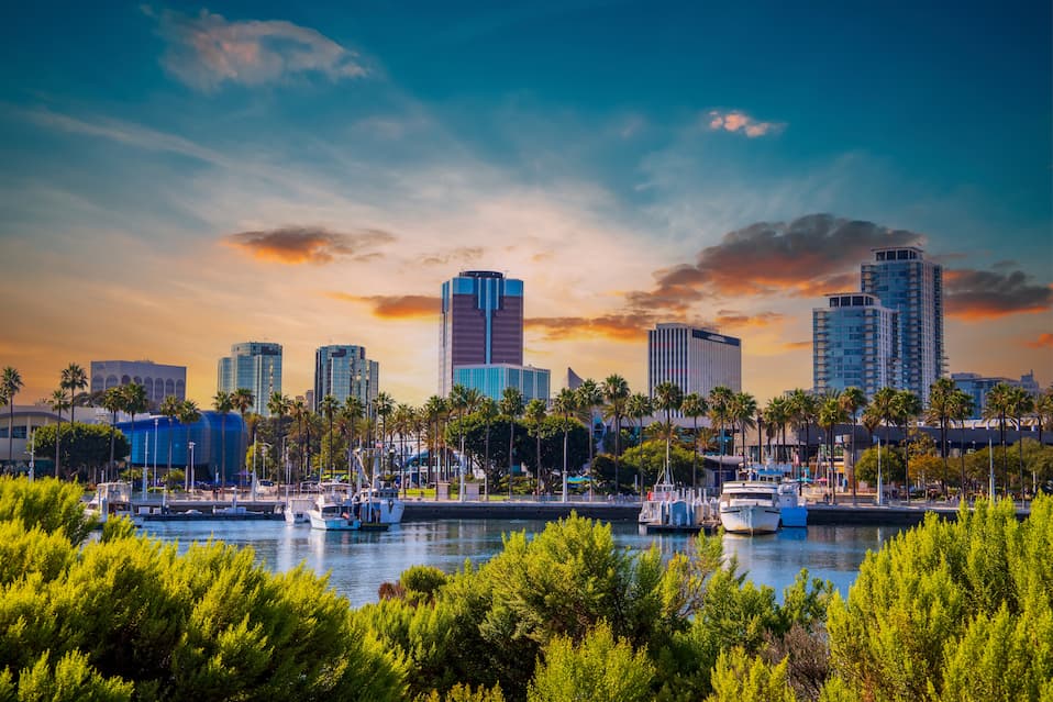 Office buildings and hotels in the city skyline with boats and yachts docked on the blue ocean water in the harbor with lush green trees and plants at sunset in Long Beach, California.
