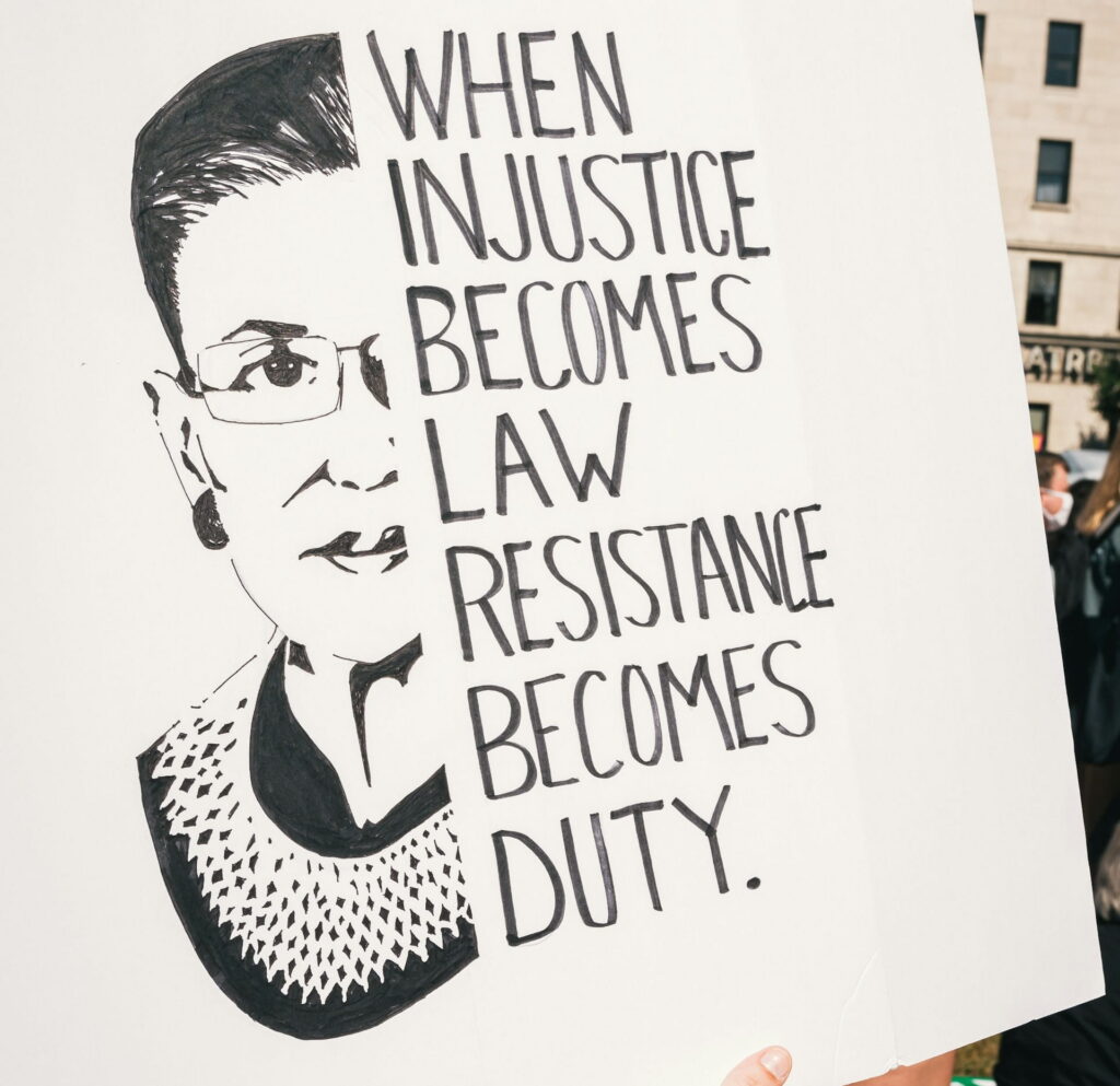 A hand-drawn protest sign held up at a public demonstration, featuring a black ink illustration of Supreme Court Justice Ruth Bader Ginsburg (1933–2020), depicted with her signature glasses and lace jabot collar. Beside the illustration, hand-lettered text reads: 'When injustice becomes law, resistance becomes duty.