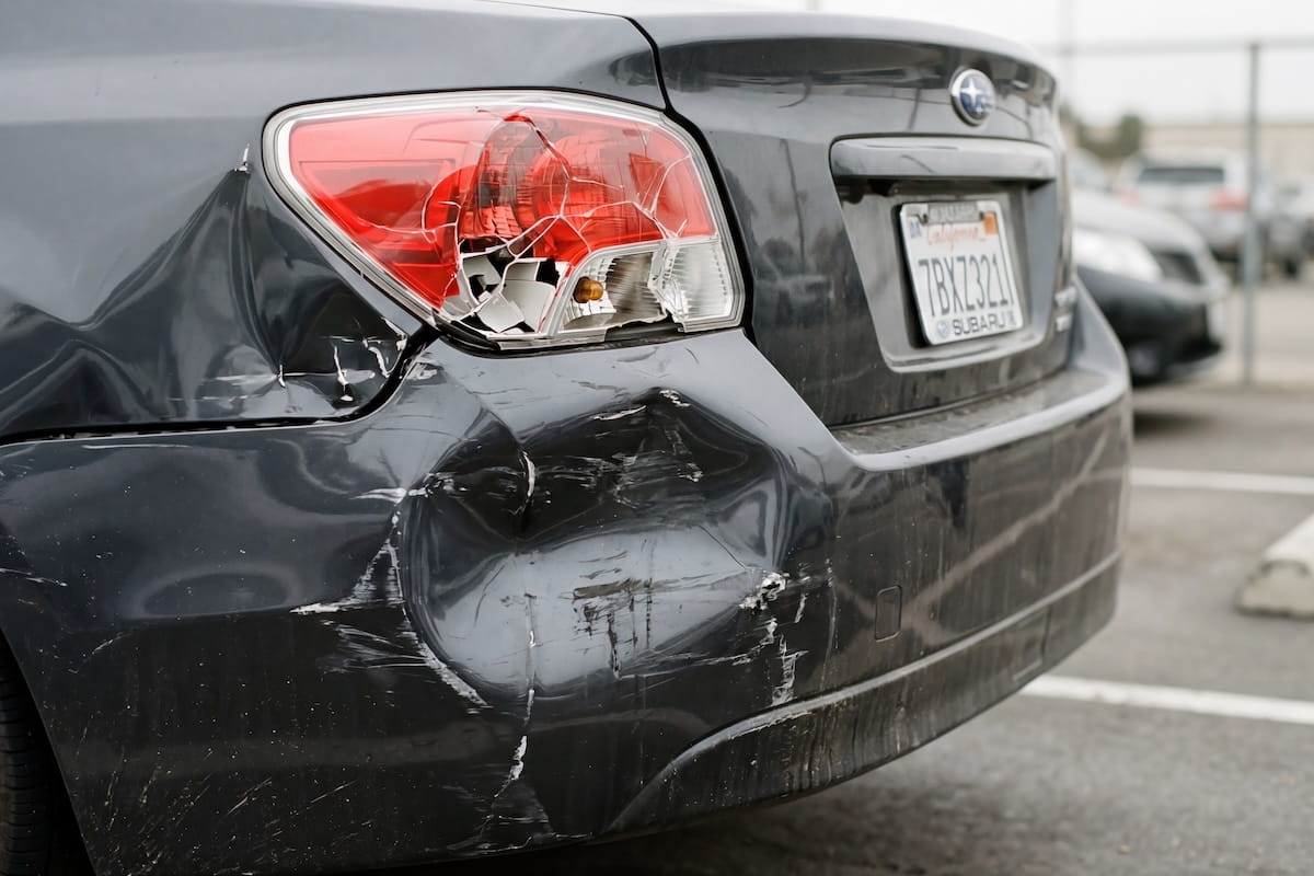 Close-up of a damaged dark gray Subaru with a heavily dented rear bumper, cracked tail light, and paint scraping consistent with a rear-end collision, photographed in a parking lot.