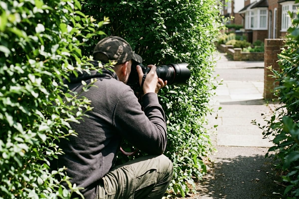 A man in a camouflage cap and gray hoodie crouches behind a dense green hedge on a residential street, aiming a professional camera with a long telephoto lens — illustrating covert surveillance tactics used by insurance company private investigators.