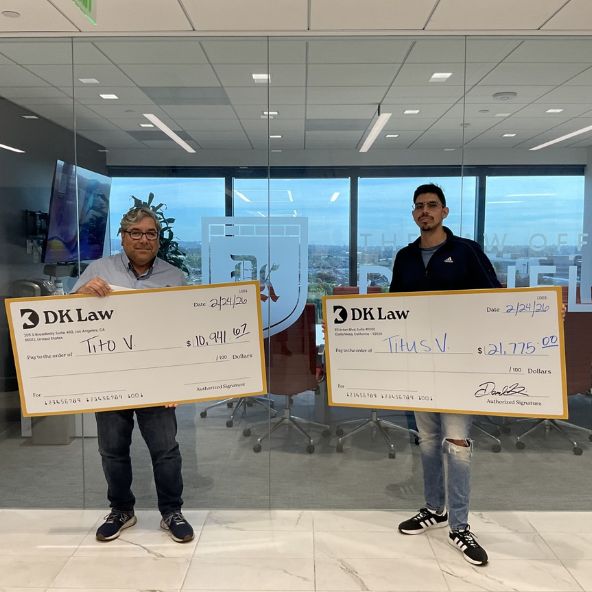 Two individuals stand in an office holding large "DK Law" checks. The check on the left is for ten thousand dollars and the one on the right is for twenty one thousand dollars.