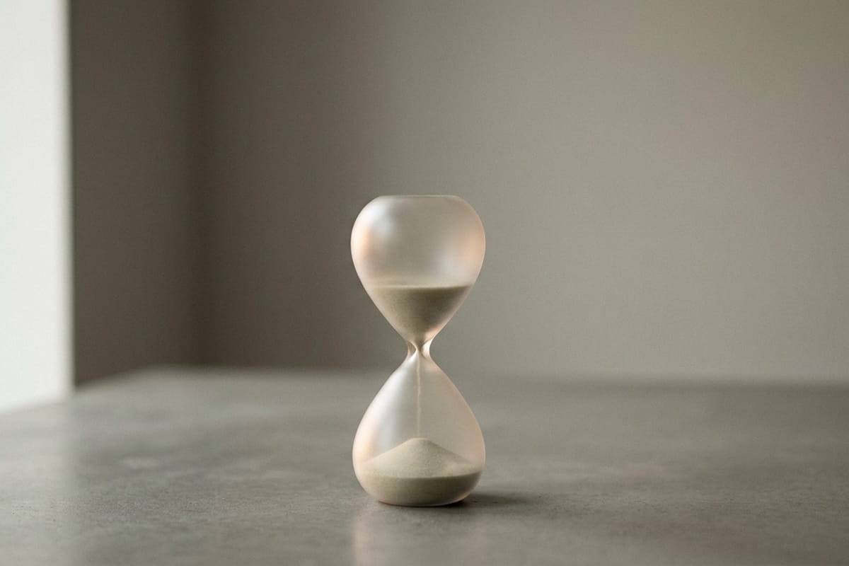 A white hourglass with sand running through it, placed on a concrete surface against a neutral gray background, symbolizing the passage of time.